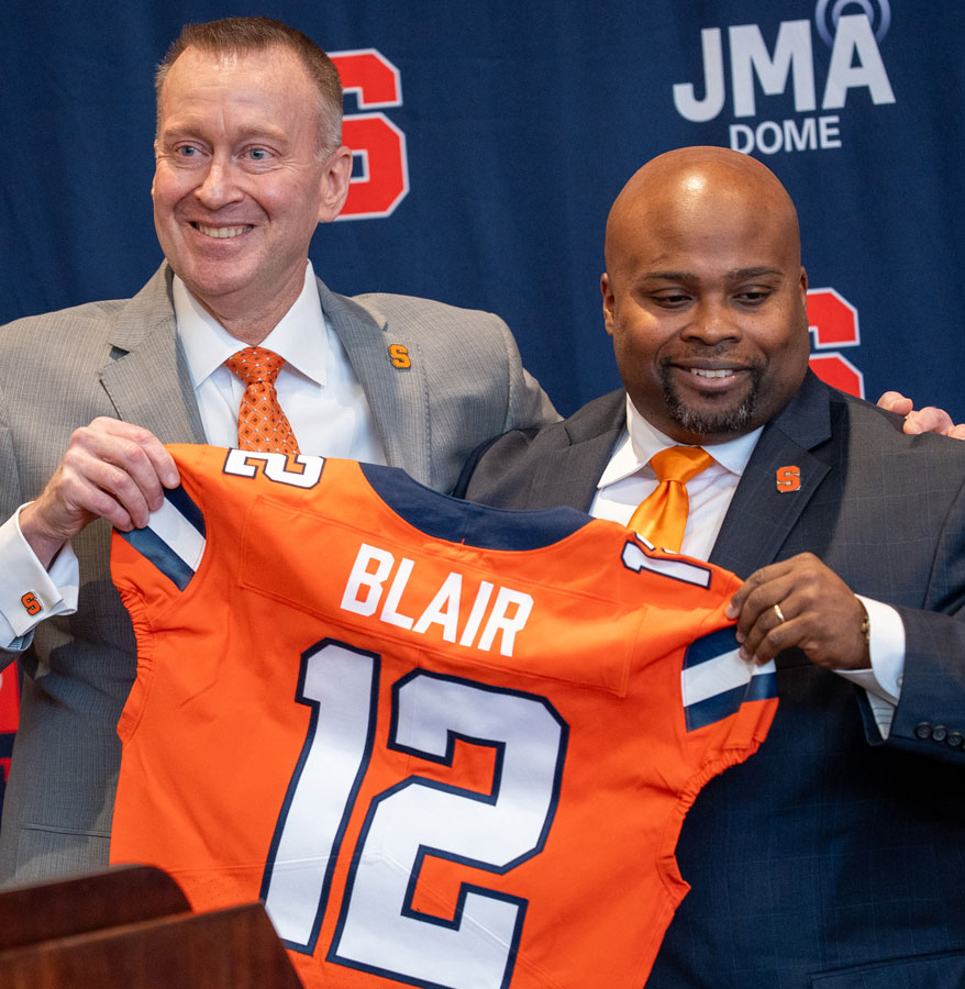 Two people stand holding an Orange number 12 Syracuse football jersey with the name Blair in white lettering on the back.