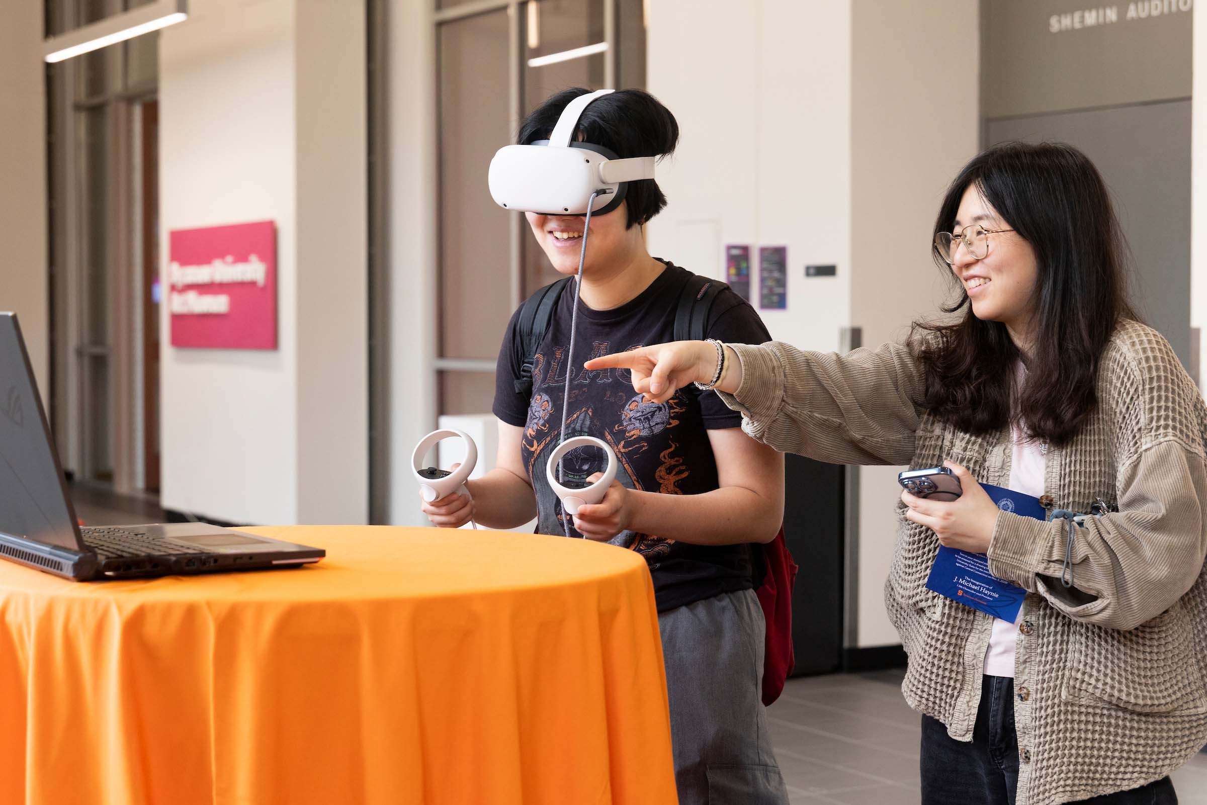 One person wears a virtual reality headset while another gestures at a computer screen. 