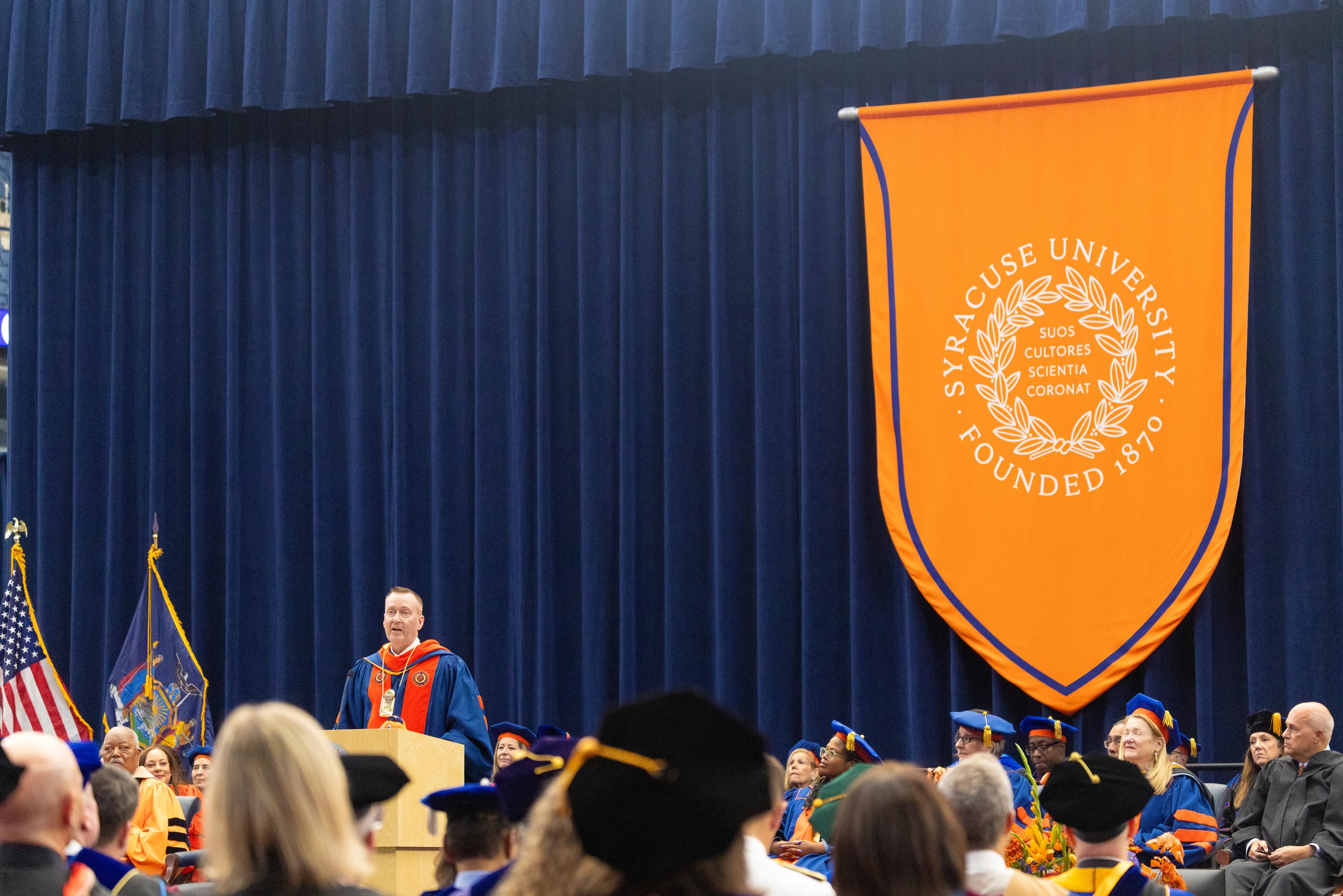 Chancellor J. Michael Haynie stands on stage in front of faculty and leaders at the university and the community. The large orange banner with the Syracuse University seal hangs to the right behind him against a blue cloth backdrop.