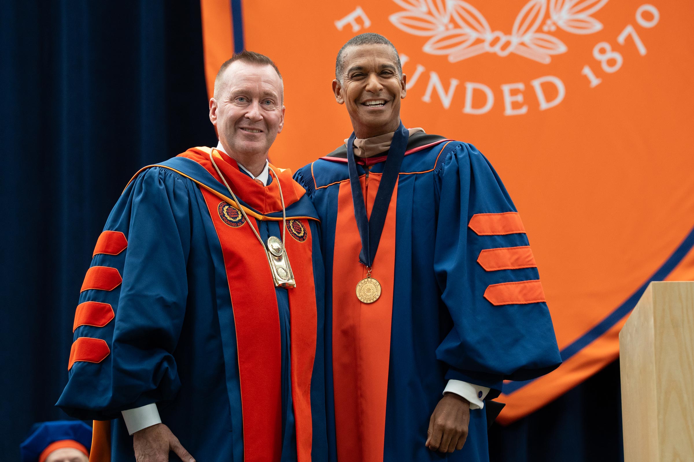 Chancellor Haynie and Board Chair Jeff Scruggs stand on stage with the large orange Syracuse banner behind them. 
