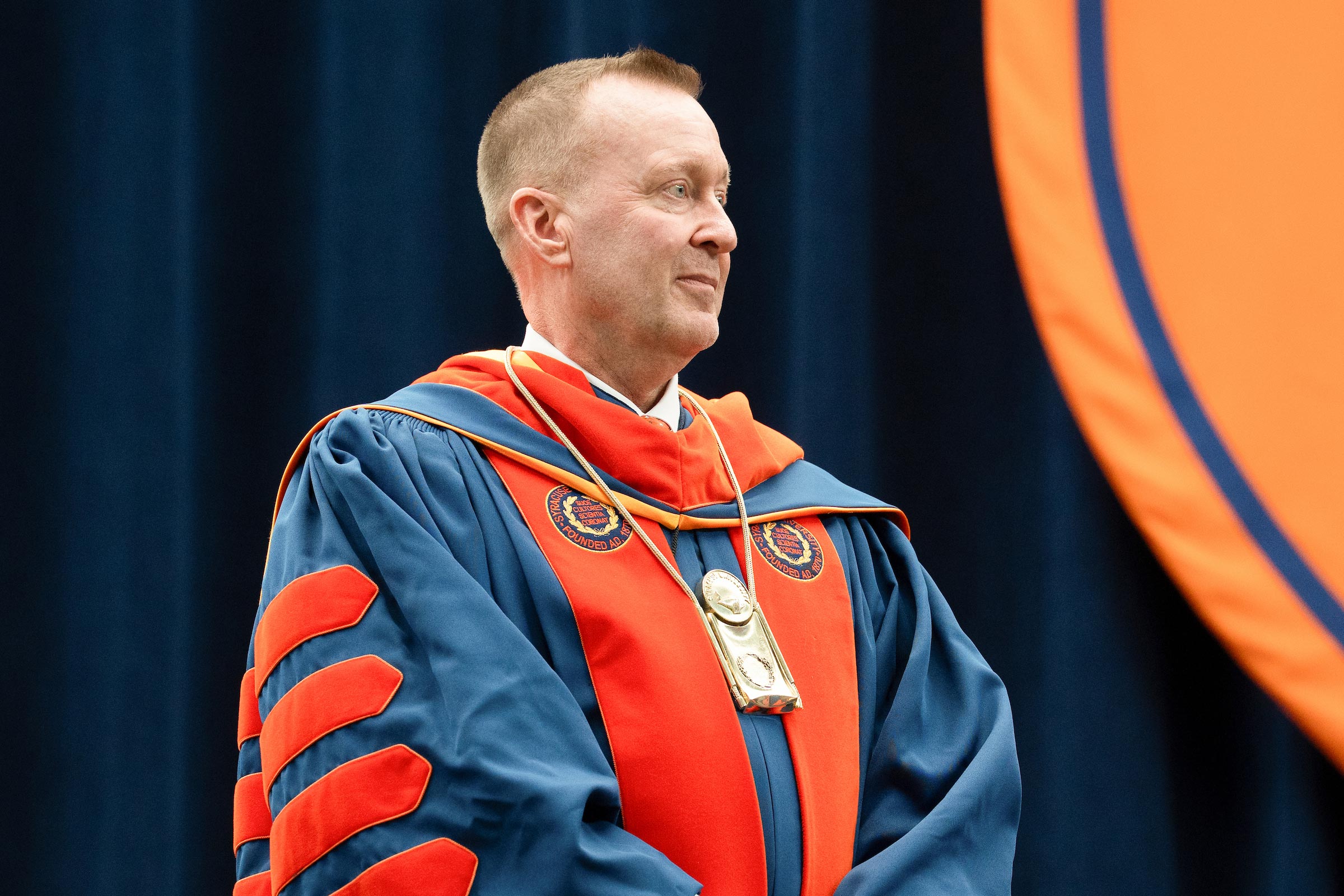 Chancellor Mike Haynie stands on stage, dressed in regalia, listening to a speaker we cannot see.