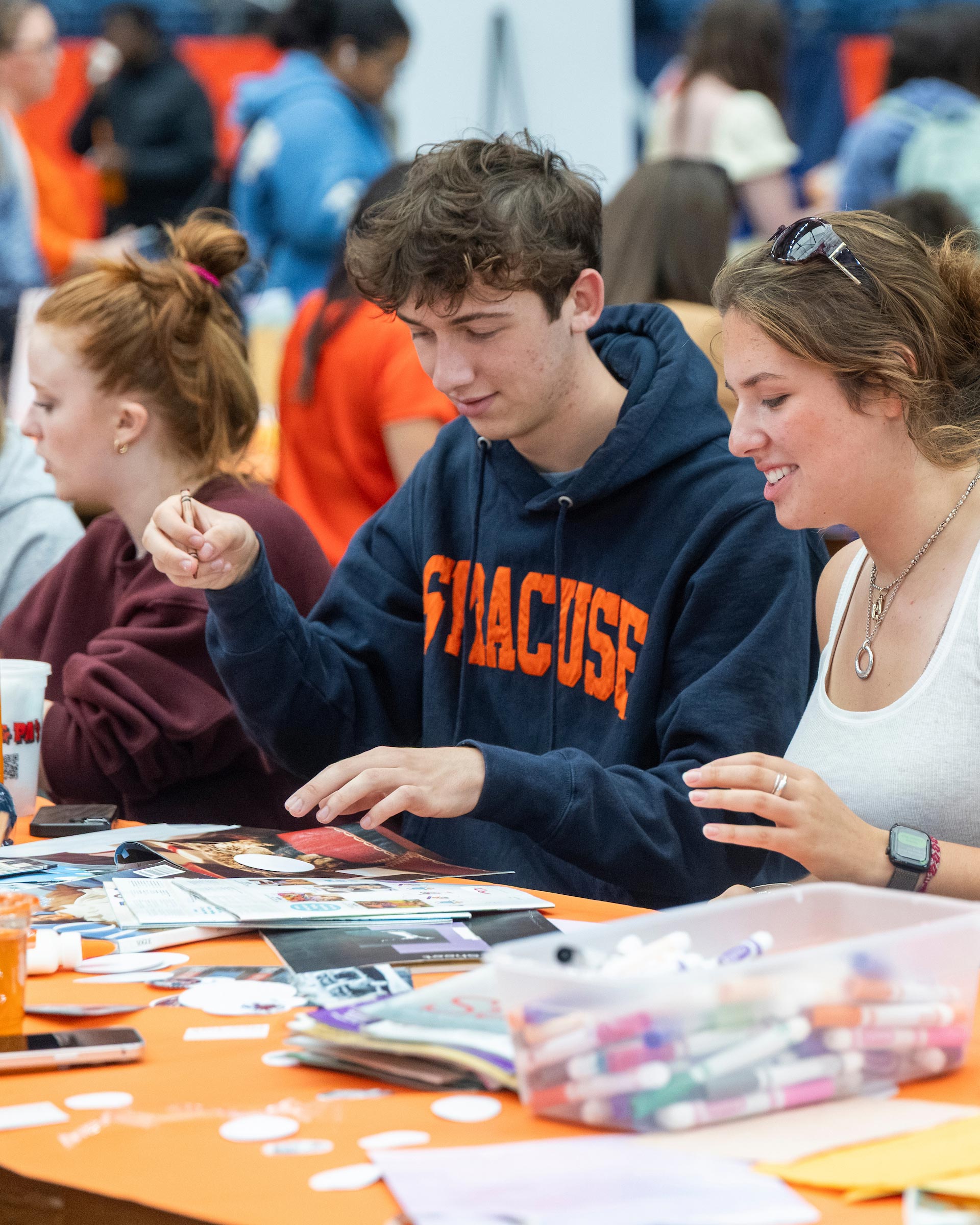 A male student wearing a blue Syracuse sweatshirt sits between two female students working on crafts at the Makerspace tables at OrangeFest. 