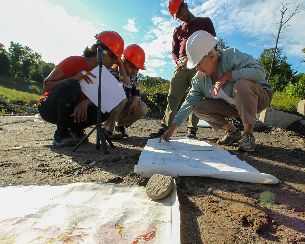 Three people squatting and looking a blue prints with person standing behind them
