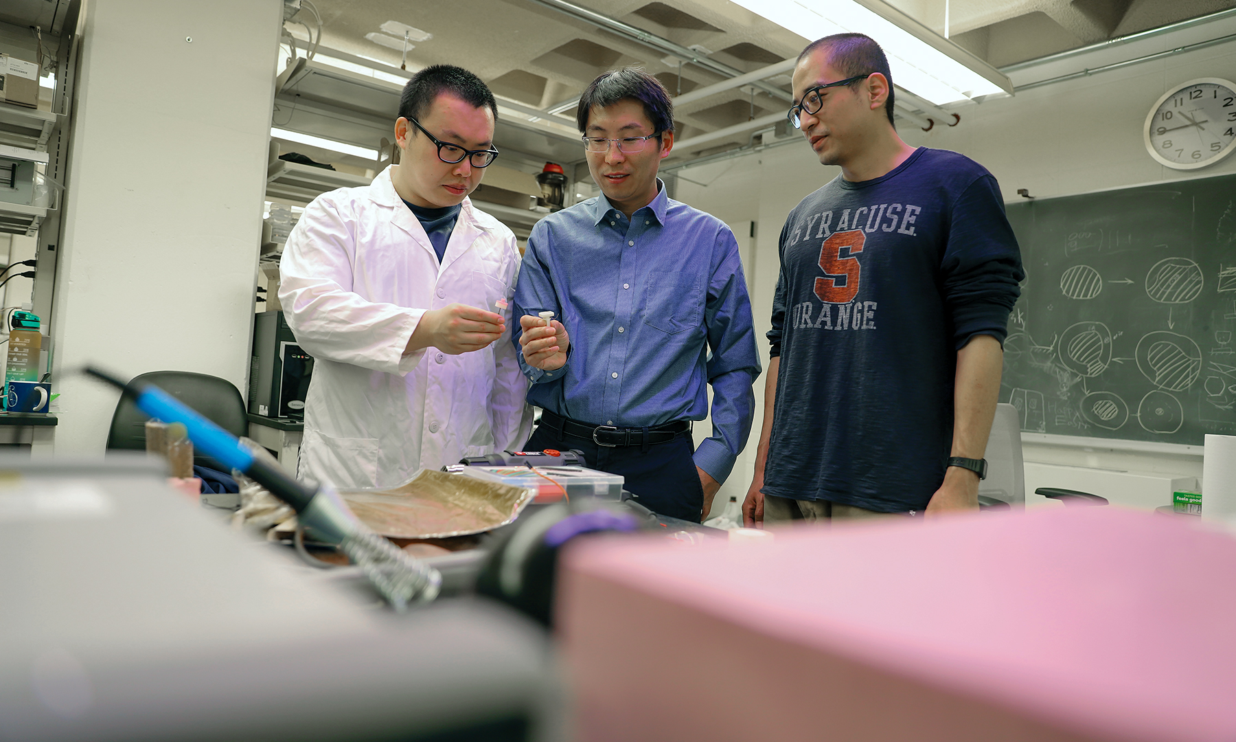 Three men standing behind a table examining a small piece of circuitry