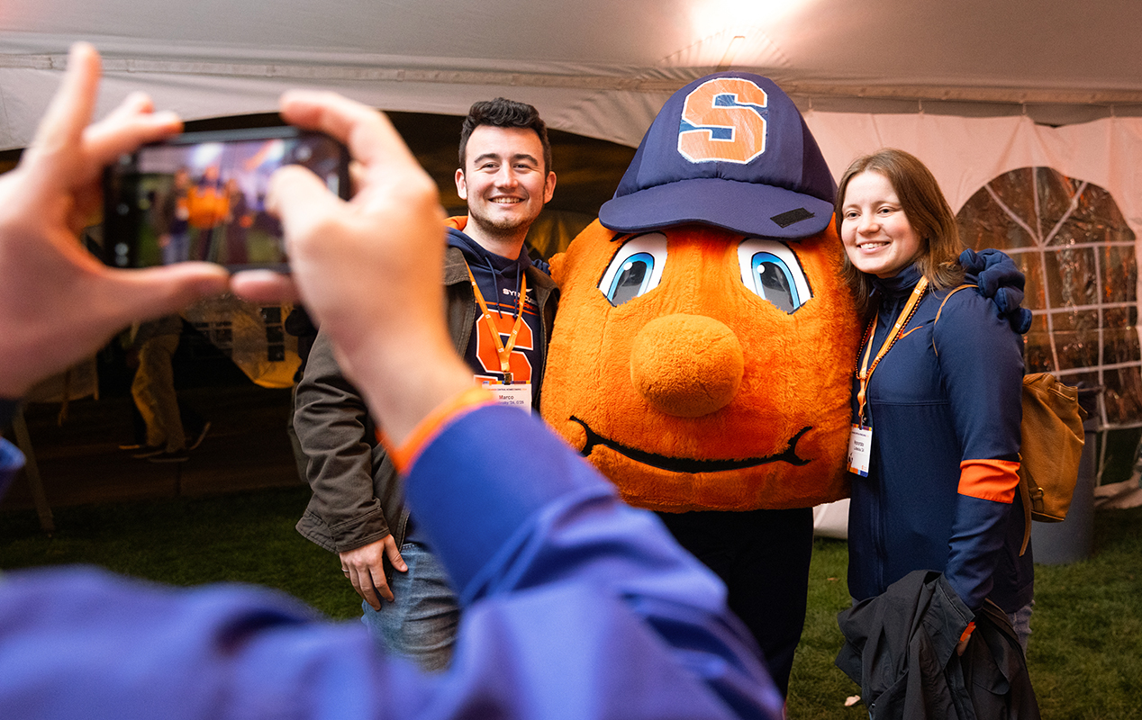A man and a woman pose with a team mascot shaped like an orange