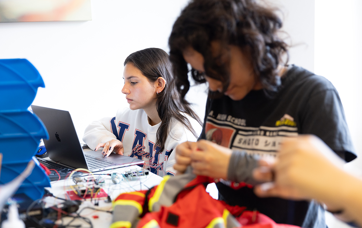 Two women sitting at a table covered in electronics looking at a laptop