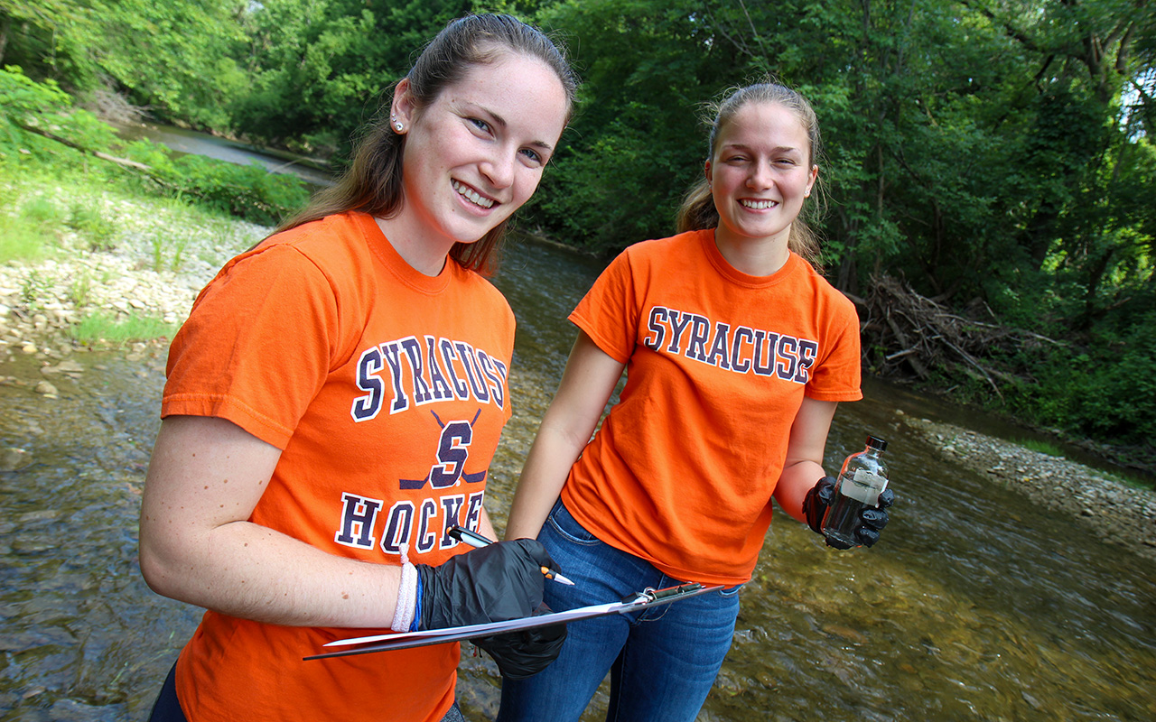 Two women standing in a stream. One holding a clipboard, and the other holding a water sample in a jar.