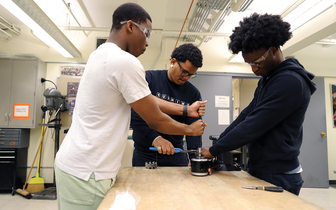 Three boys standing over a table using a screwdriver 