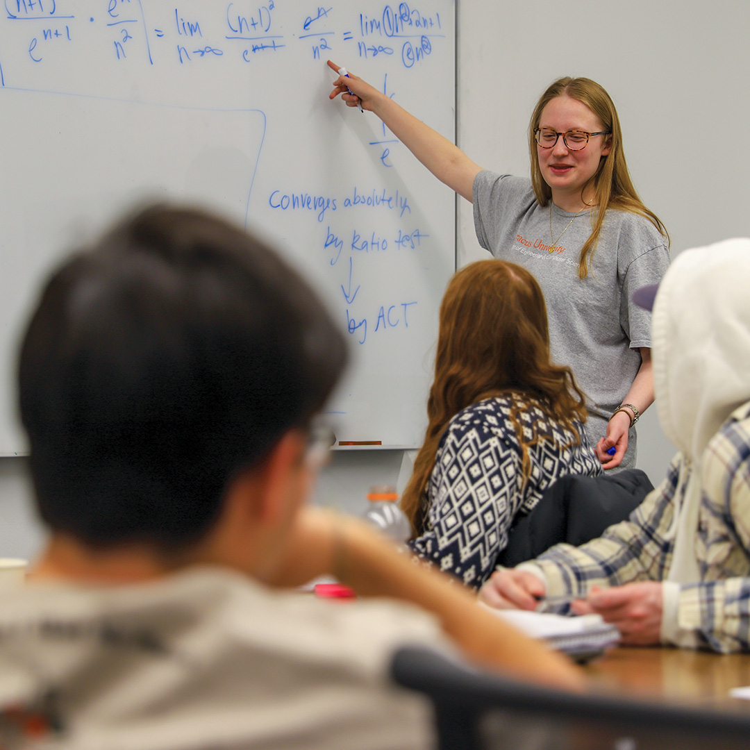 A girl pointing at a whiteboard with math written on it and talking to three students.