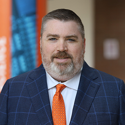 A man with short hair, a beard, and a blue suit with an orange tie.