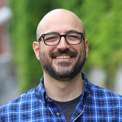 A man with no hair, a black beard, wearing glasses and a blue checkered casual shirt.