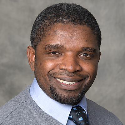 A man with dark short hair. He is smiling and wearing a grey sweater over a light blue dress shirt and tie.