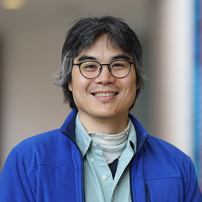 A man wearing glasses, and a green shirt with a royal blue jacket over it.