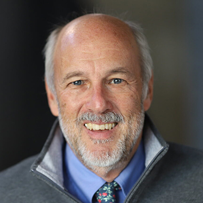 A man with no hair, a grey beard, and wearing a grey sweater over a blue dress shirt.