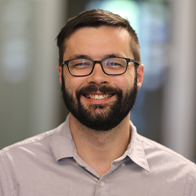 A man with dark hair and a beard, smiling, and wearing glasses with a grey collard shirt.