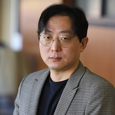 A man with dark hair. He is wearing glasses and grey blazer over a dark shirt.