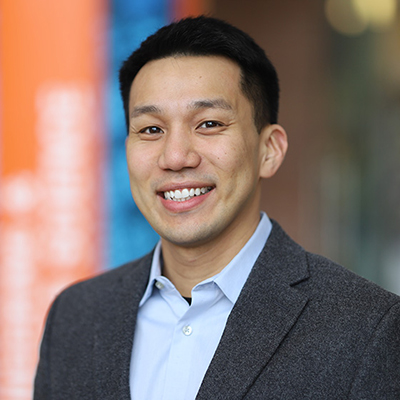A young man with dark hair. He is smiling and wearing a dark blazer or a light blue shirt.