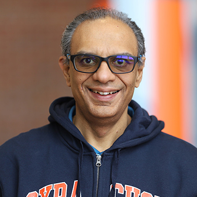 A man with no hair, wearing tinted glasses and zip up Syracuse hooded sweatshirt.