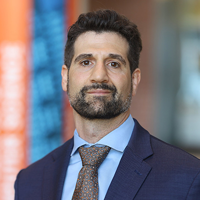 A man with dark hair and beard, and wearing and navy blue suit with a brown dotted pattern tie.