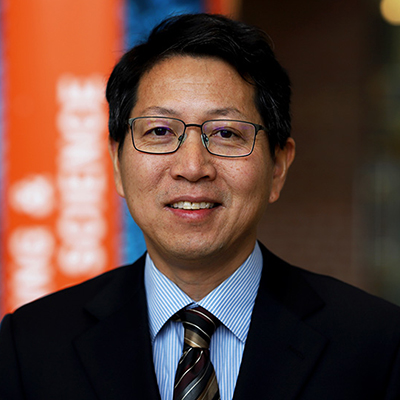 A man with dark hair smiling. He is wearing glasses and dark suit with a light blue shirt.