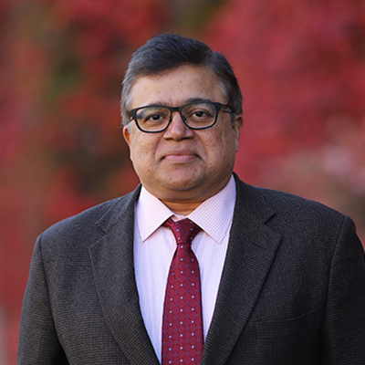 A man smiling, and wearing thick black rim glasses, a dark suit, white shirt, and red tie.