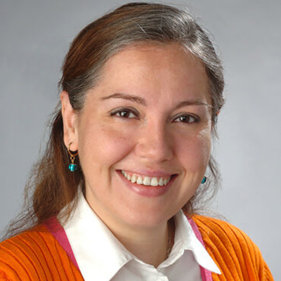 A woman with light brown hair smiling. She is wearing a white blouse with an orange cardigan. 