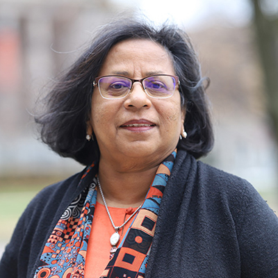  A woman with medium length dark hair. She is wearing glasses, a dark color sweater over a multi-colored top.