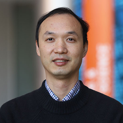 A man with short dark hair wearing a black sweater over a checkered dress shirt.
