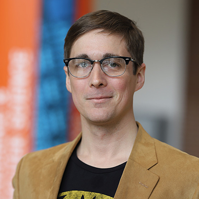 A man with short brown hair smirking. He is wearing glasses, and a brown blazer over a black tee shirt.