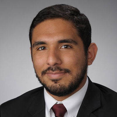 A man with dark hair and a beard. He is wearing a dark suit with a white shirt and red tie.