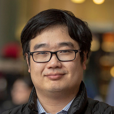 A man with dark hair, smirking and wearing glasses.
