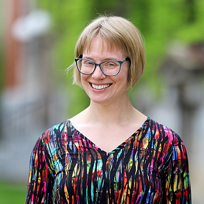 A woman wearing glasses and smiling. She is wearing multicolored shirt.