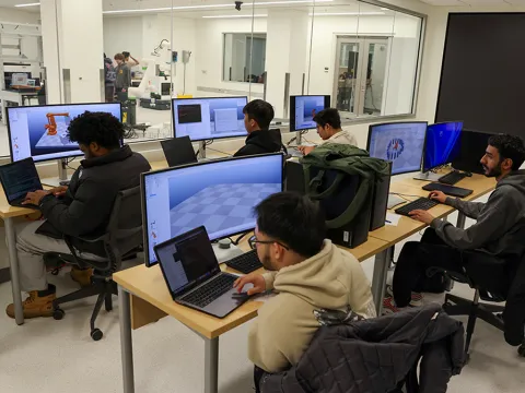 Five students sitting at tables and looking at monitors in a computer lab