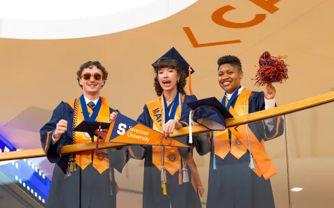 On man and two women wearing graduation regalia and standing against a railing. 