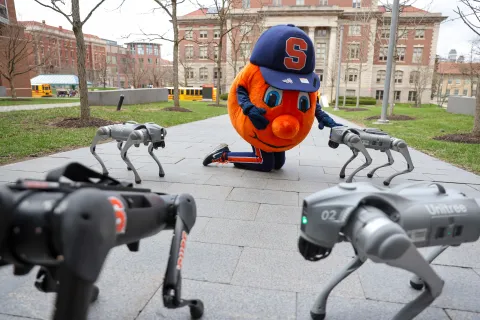 An orange mascot pretending to pet a robot dog. Surrounded by three additional robot dogs. 