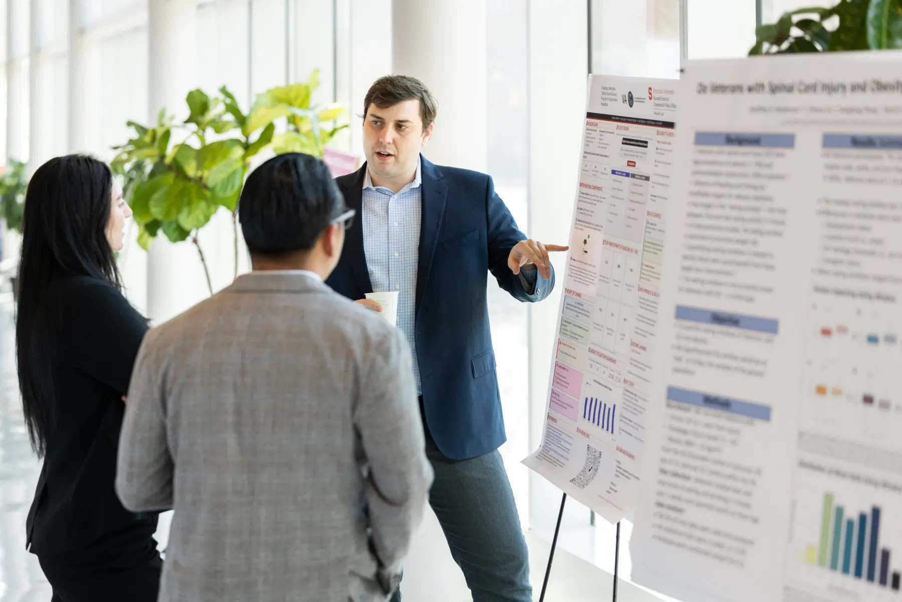 A man standing beside a research poster points at a data point while talking to a woman and man standing in front of the poster.