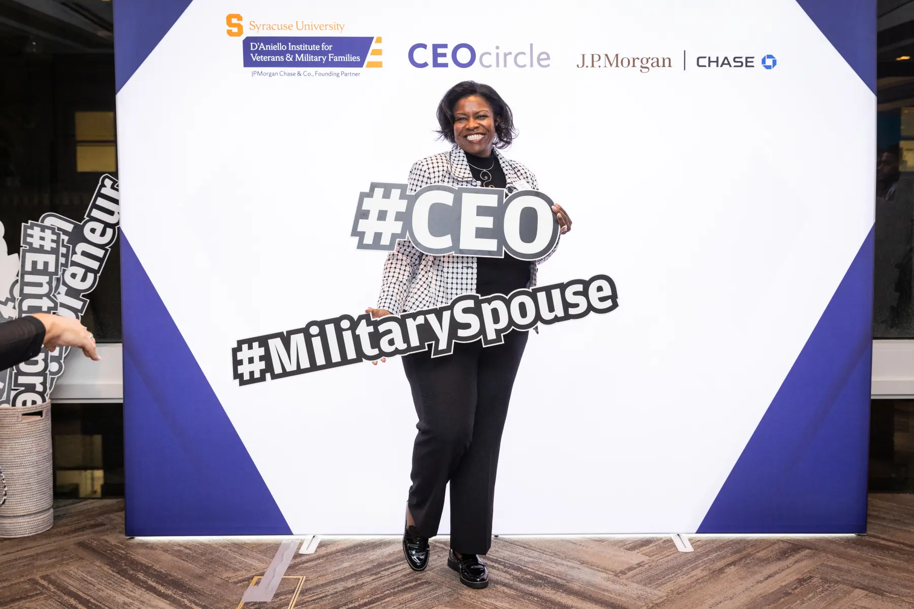 A woman poses for a photo holding two prop signs reading "#CEO" and "#Military Spouse" in front of a Syracuse IVMF CEOCircle JP Morgan Chase wall. 