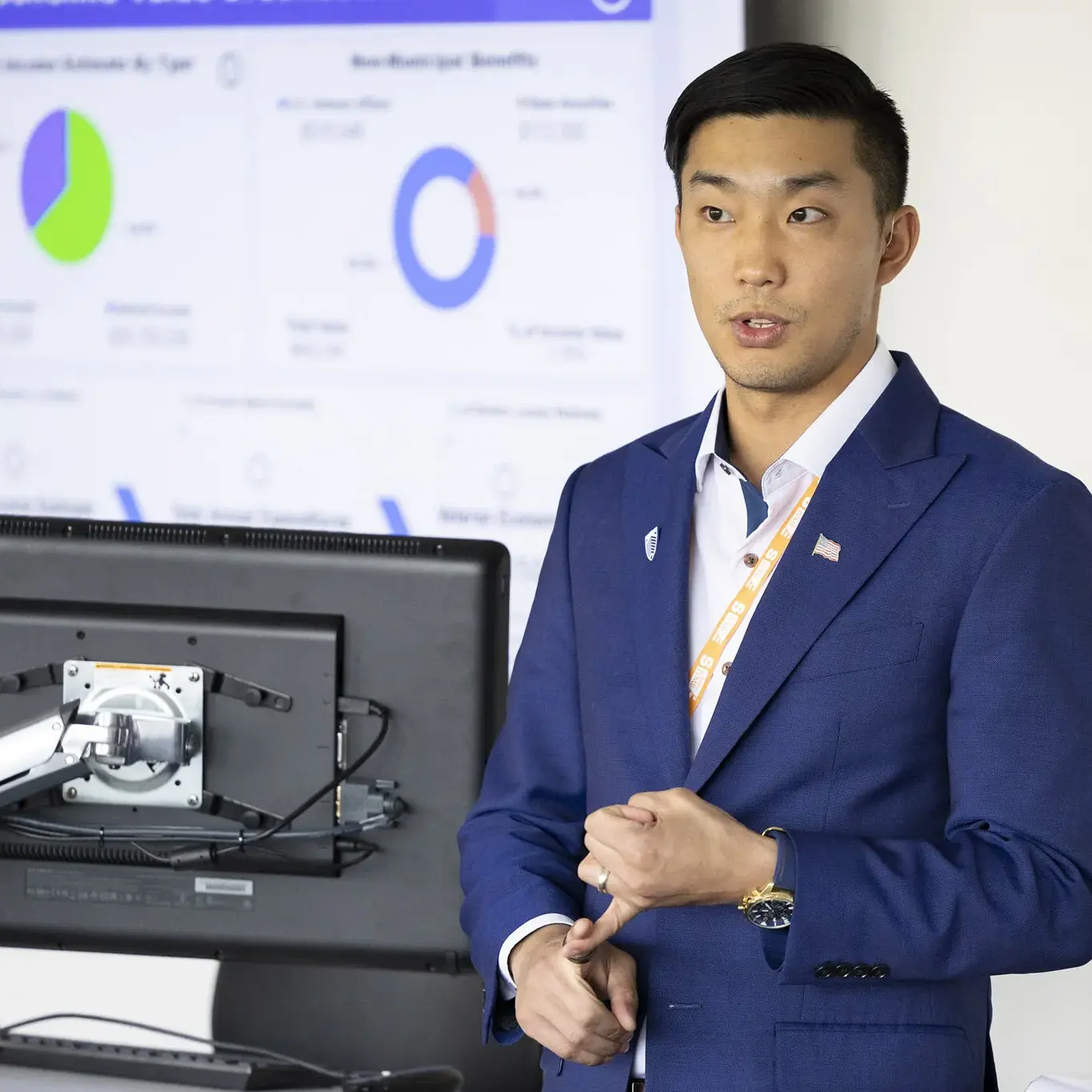 A man wearing a suit addresses an audience while standing to the side of a screen displaying charts and graphs at the front of a conference room.