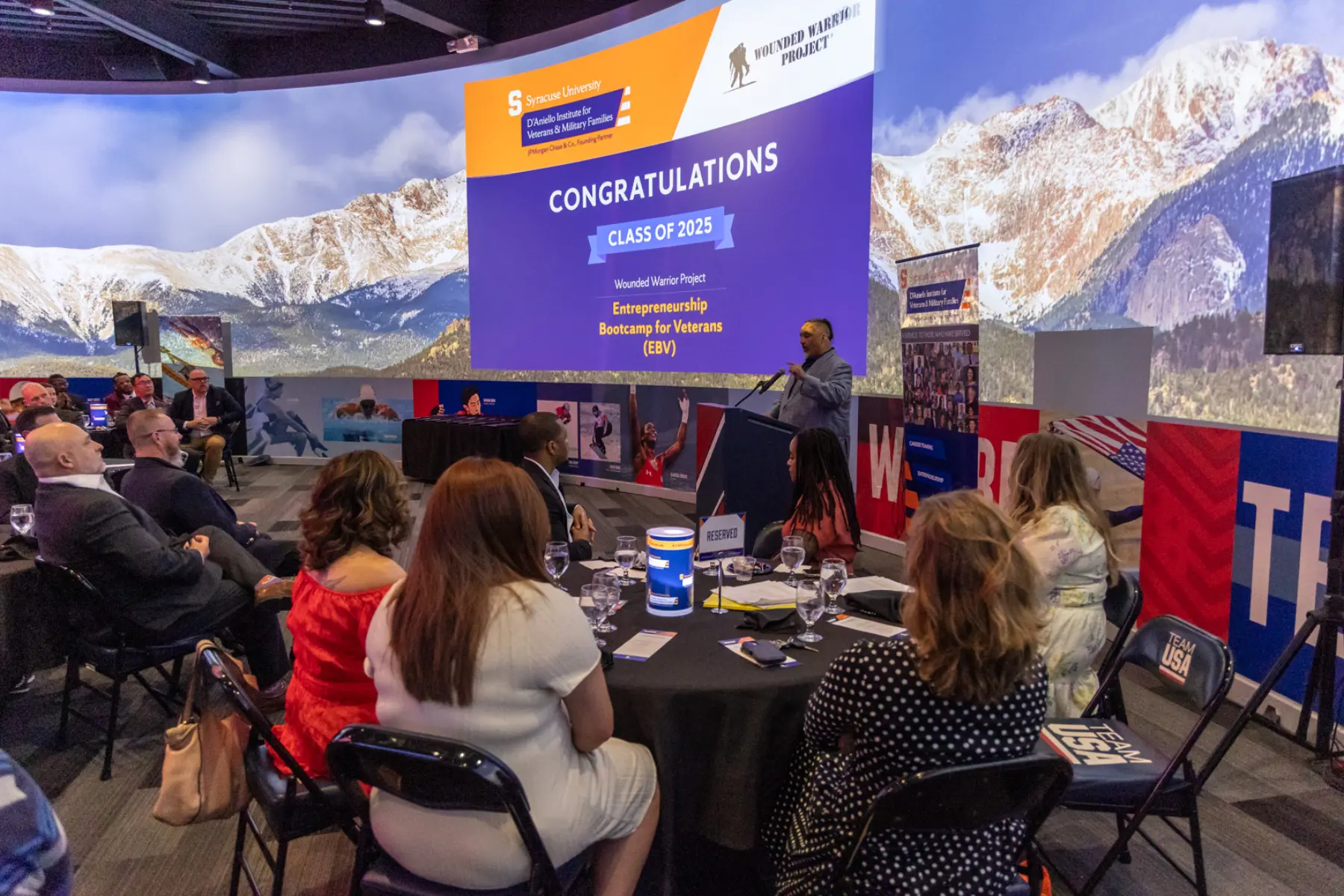 A speaker behind a podium addresses the EVB audience with a large mountainscape projected on the curved wall behind him.