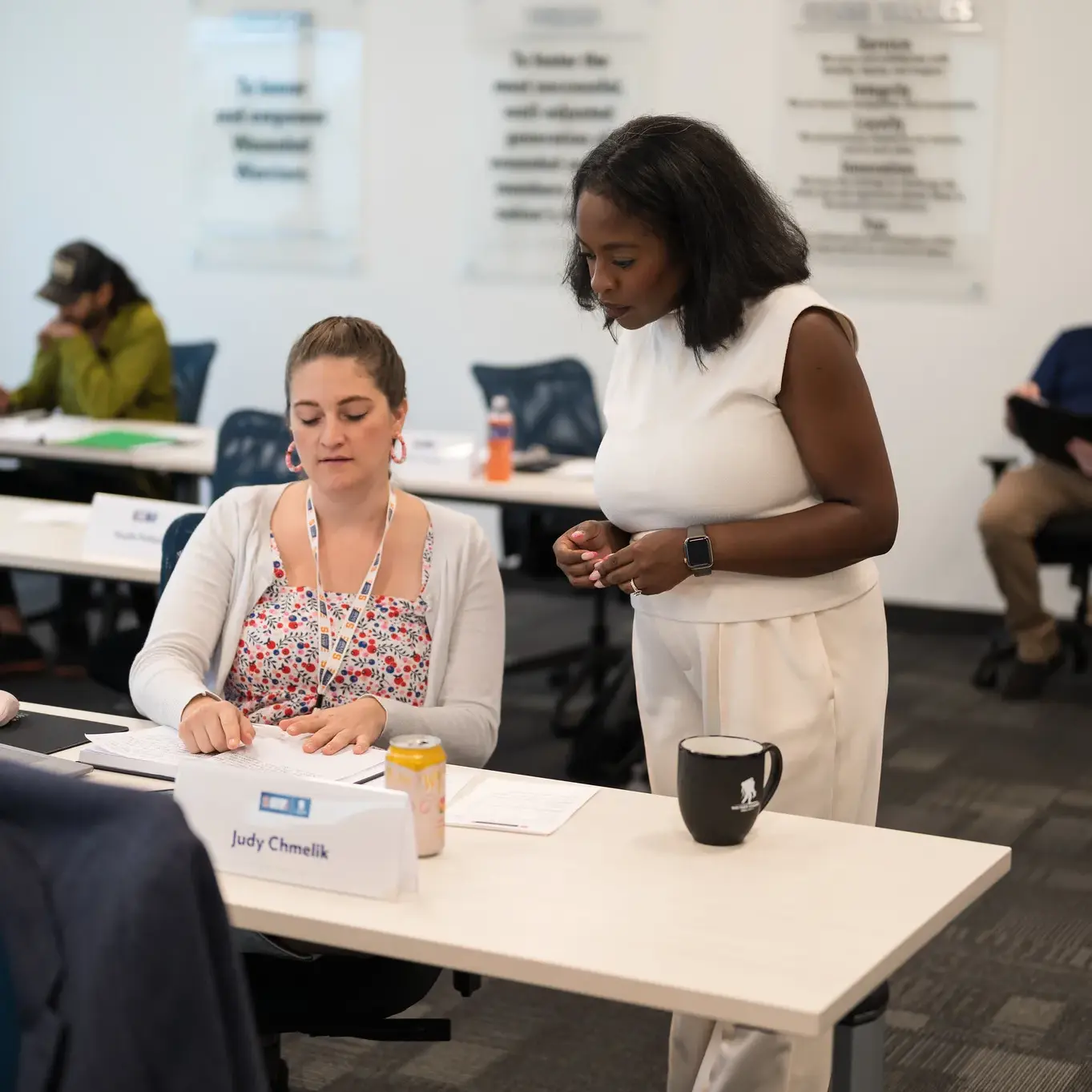 In a classroom, a woman stands beside a seated attendee. Both are looking at an open notebook on the table.