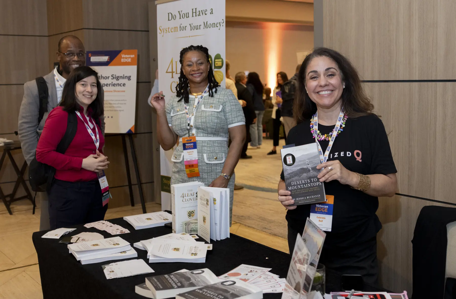 Three women and a man standing in a lobby area smile for a photo. A women standing behind a table proudly holds one of the books on her table.