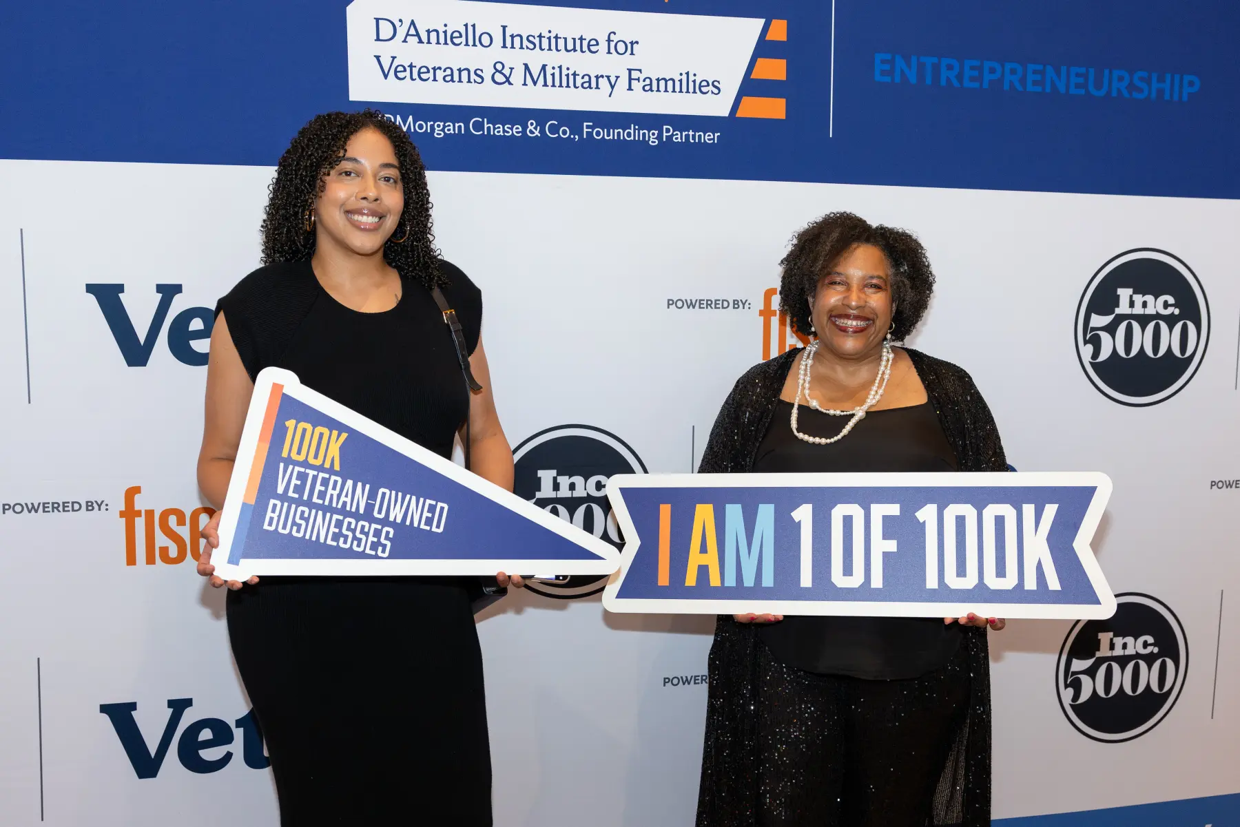 Two women pose together in front of a Syracuse IVMF Entrepreneureship step and repeat wall while each holds a sign with "100K"