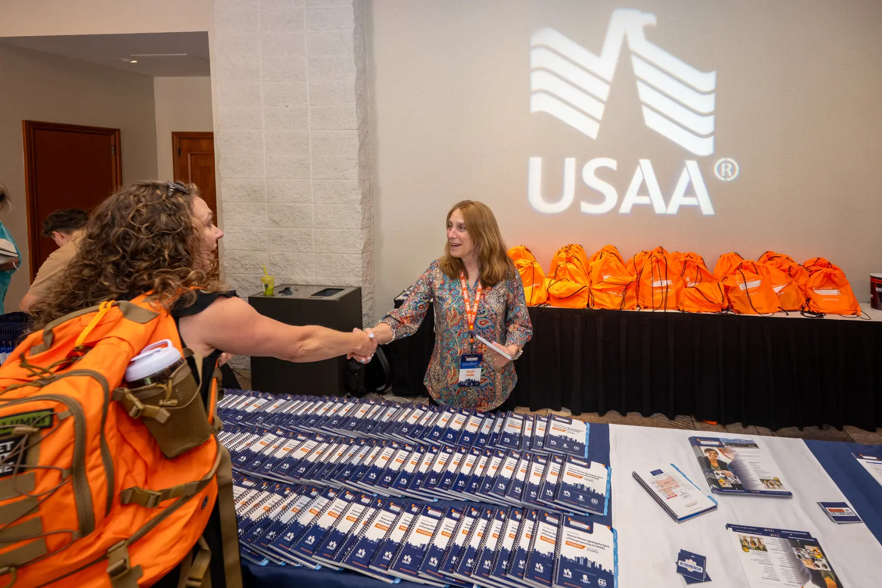 An IVMF staff member greets an attendee from behind a table covered with program books and other IVM mateirals. A USAA logo is projected on the wall.