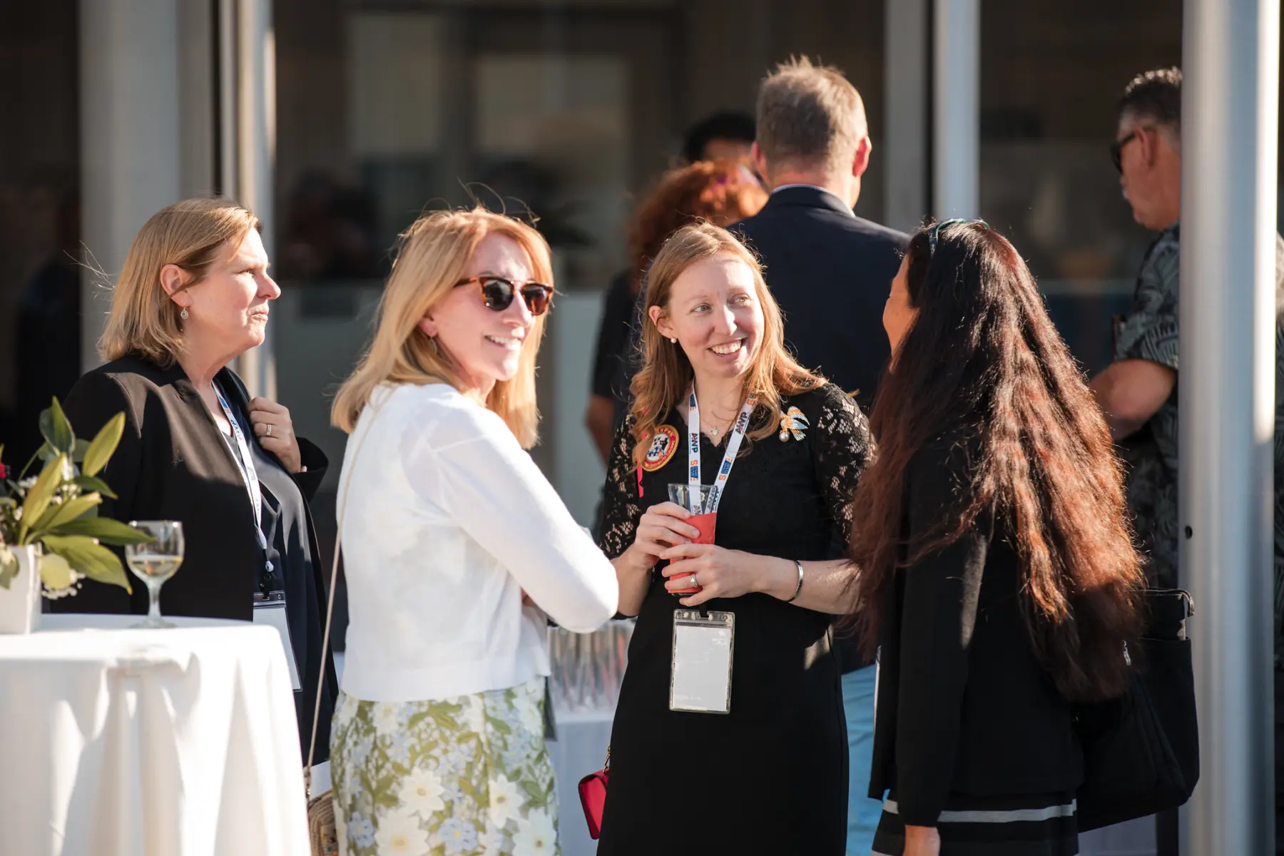 In a sunny outdoor patio setting, three women engage in light conversation and laugh while others move around about them.