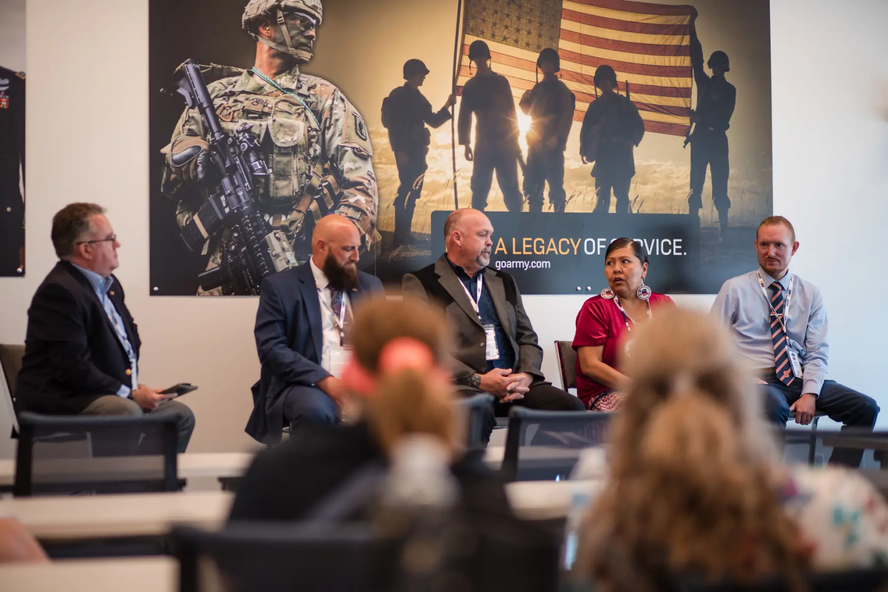 A moderator and four panelists sit at the front of a conference room with a large "Legacy of Service" mural to the backs. A woman panelist is speaking.