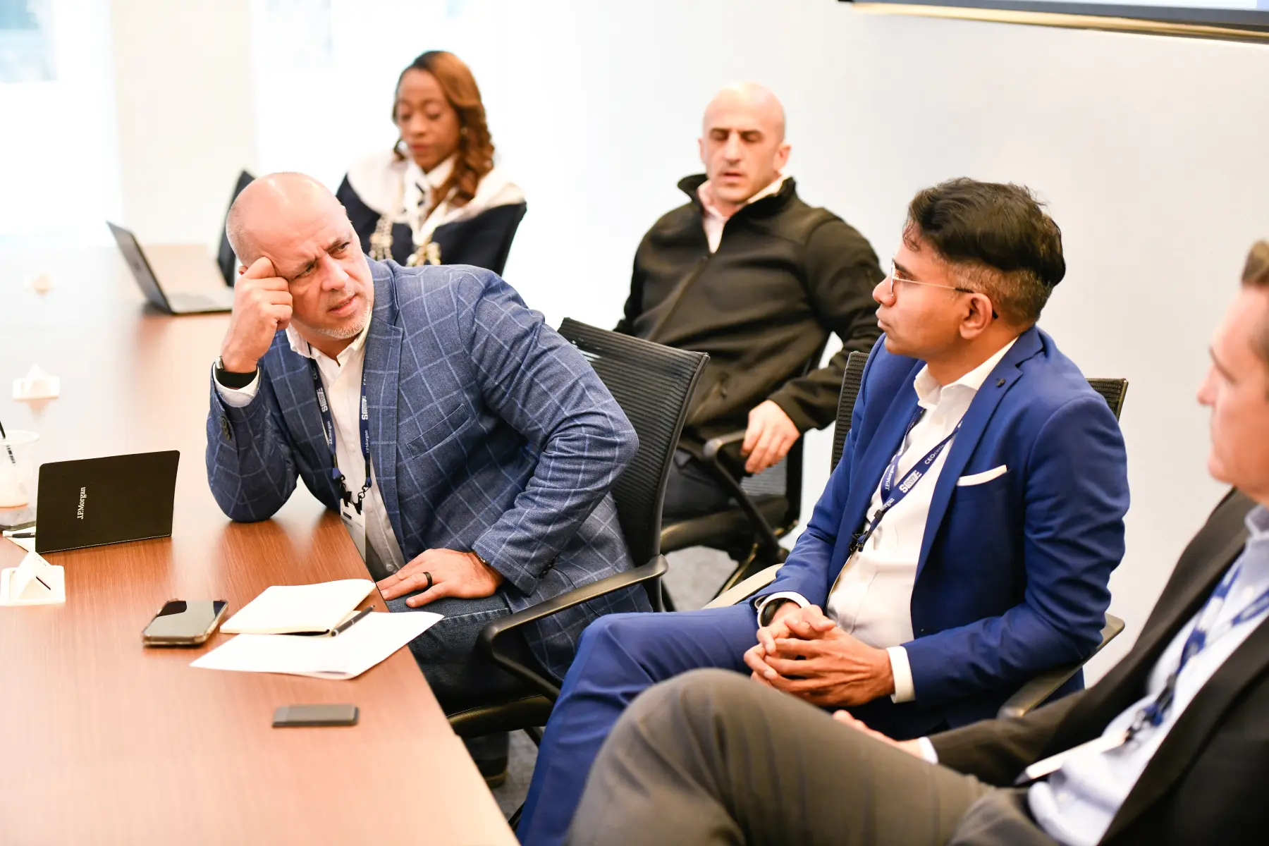 Two men stting beside one another at a conference table have a serious conversation with one leaning on table looking at the other.