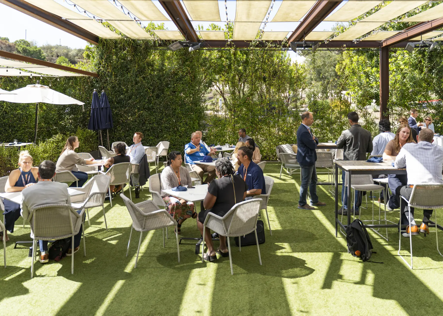 Event attendees sit in small groups and talk during a break in sessions, enjoying a shaded tree-lined patio setting.