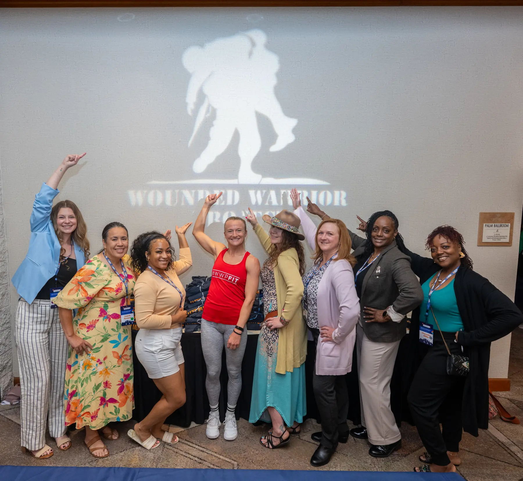 Eight women pose together smiling and pointing toward a large projection of the Wounded Warrior Project logo on the wall behind them.