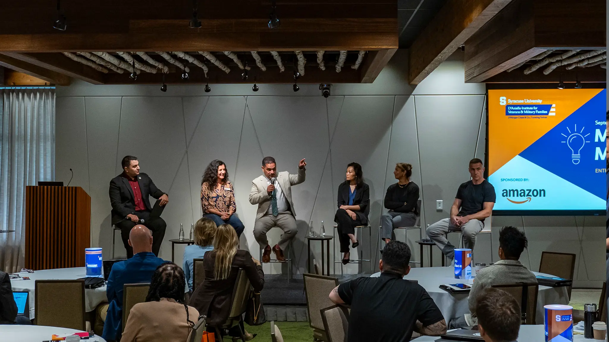 A panel of 5 speakers and a host sit on a spotlight lit stage. The panelist in the middle is speaking while in the spotlight.  