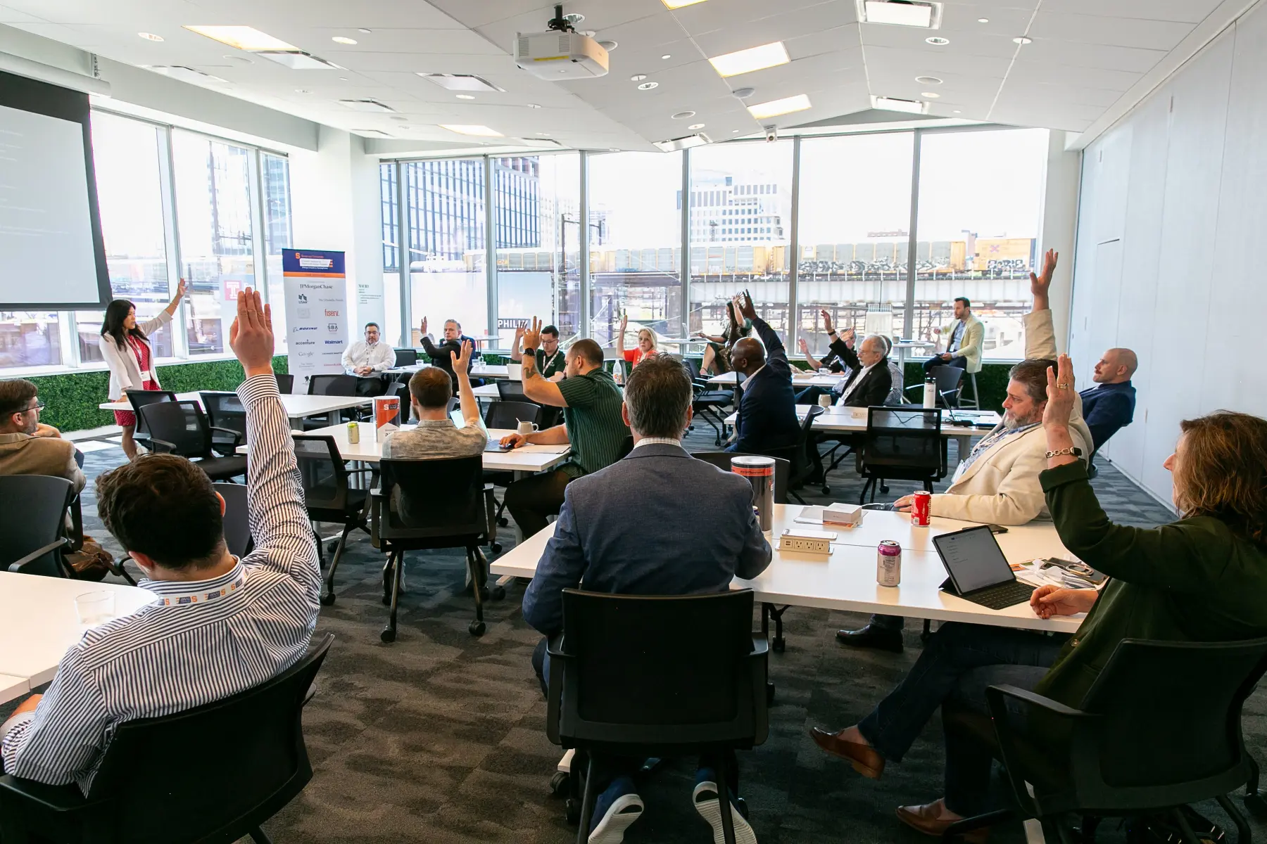 A large brightly lit conference room is filled with MEF attendees raising their hands and looking toward the presenter who also has her hand raised.